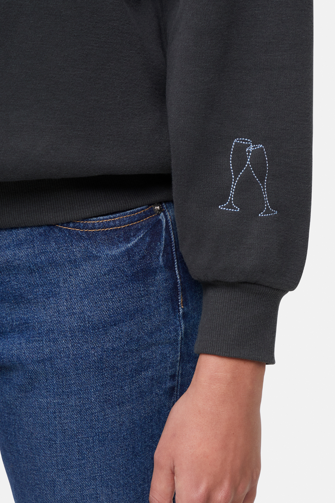 Close up of black sweatshirt left sleeve hem with with a 2 small embroidered champagne glasses graphic worn by a woman, paired with blue jeans