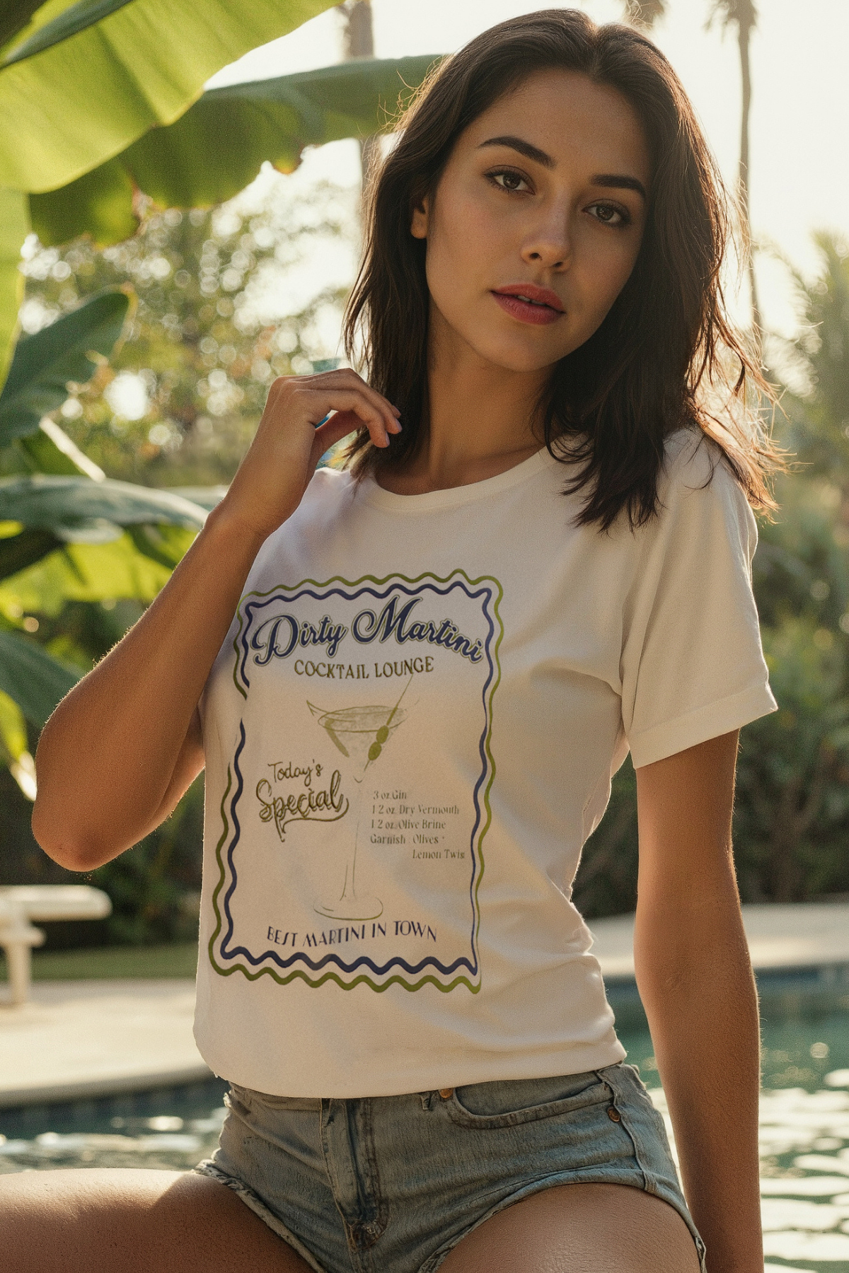 A woman wearing a white crop tee with a graphic that includes text and a martini illustration, paired with denim shorts, standing near a pool.