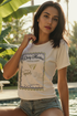 A woman wearing a white crop tee with a graphic that includes text and a martini illustration, paired with denim shorts, standing near a pool.