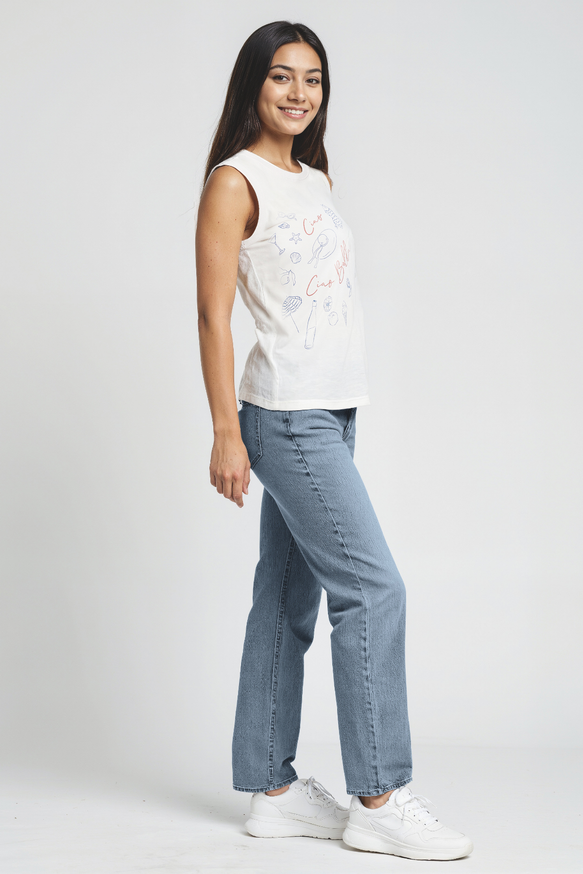 Woman wearing a white sleeveless top with colorful designs and blue jeans on a white background