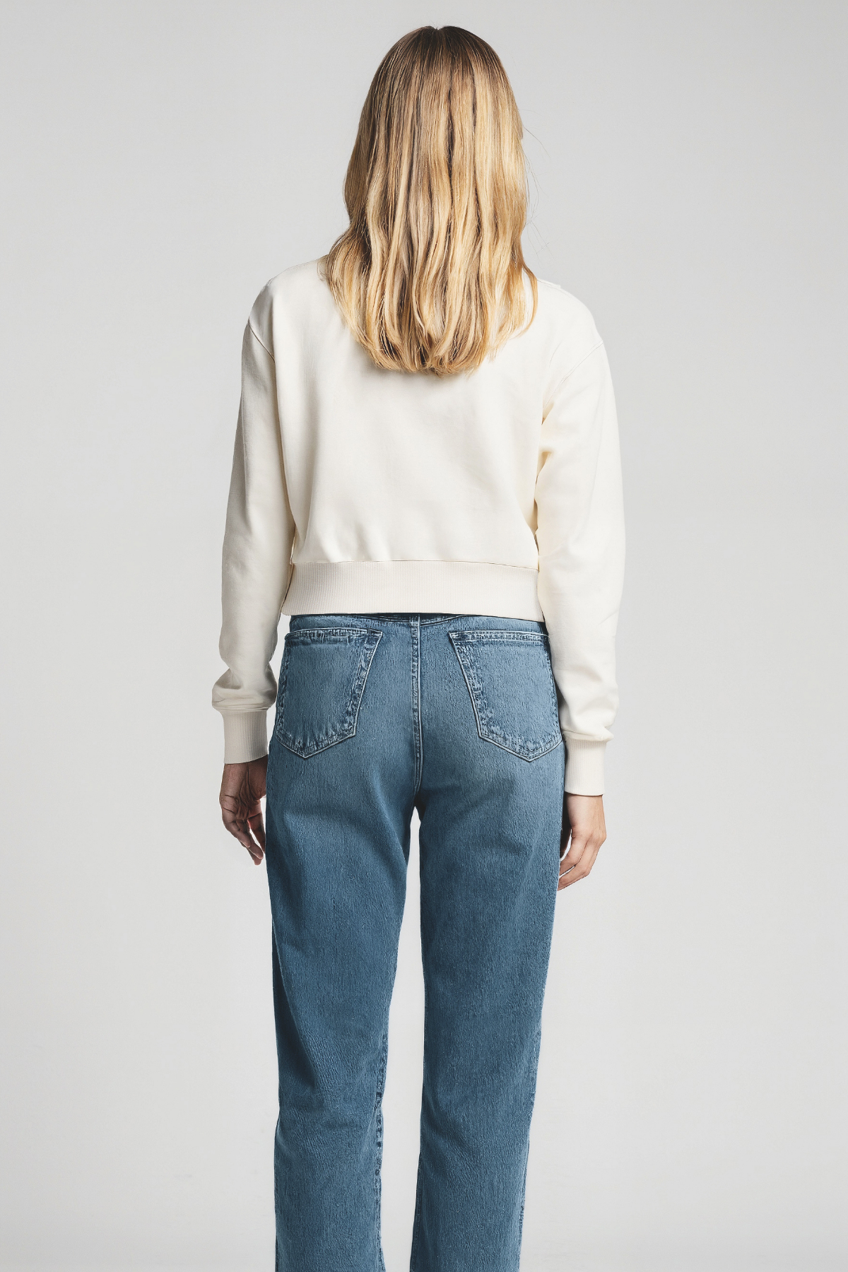 Back of women wearing a white sweater and blue jeans on a plain background