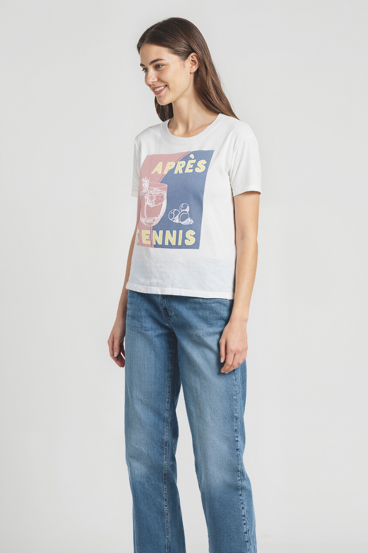 Woman wearing a white t-shirt with colorful cocktail graphic design and "APRES TENNIS" text with blue jeans on a plain background