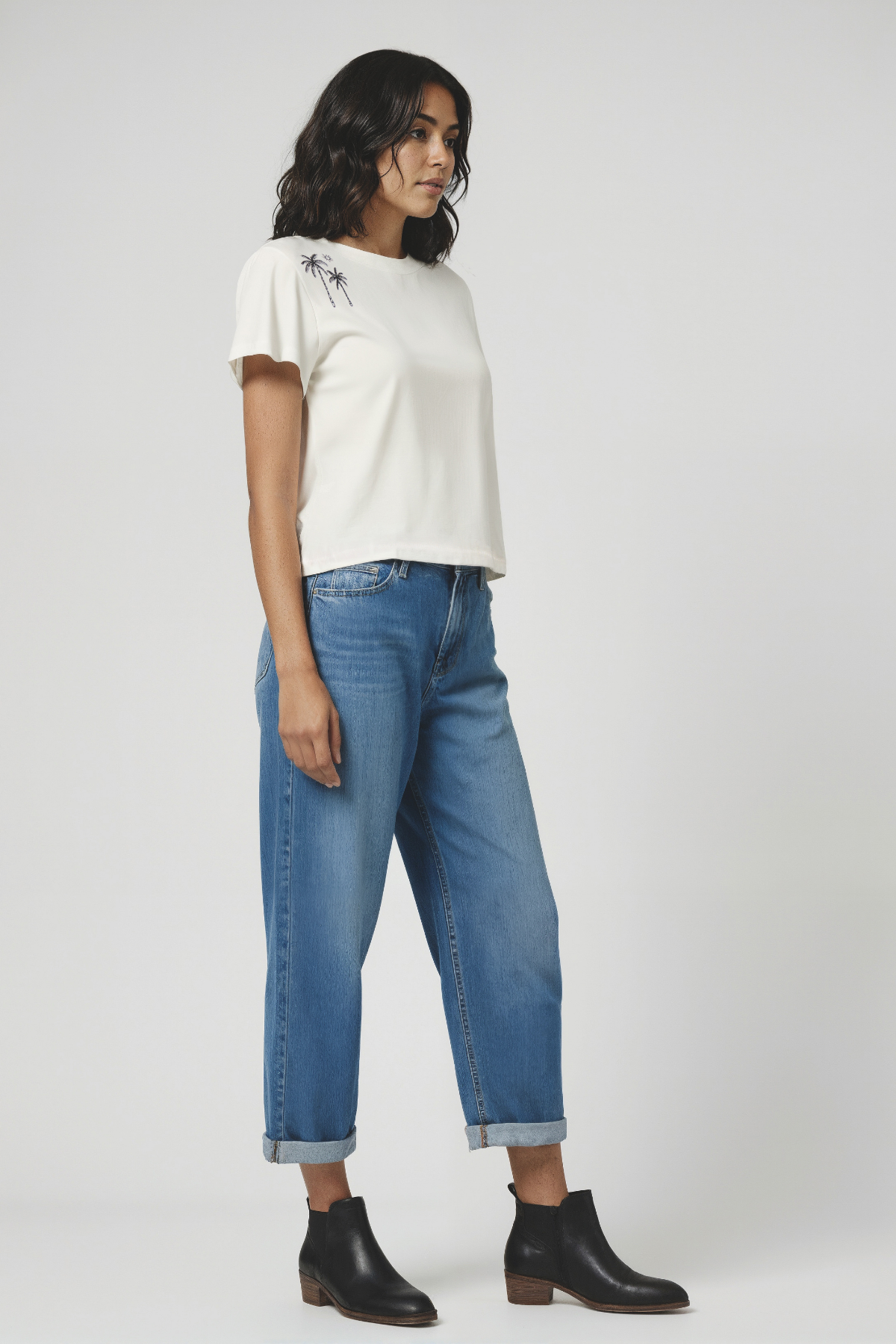 Woman wearing a white t-shirt with a palm tree design on top right shoulder and blue jeans on a plain background