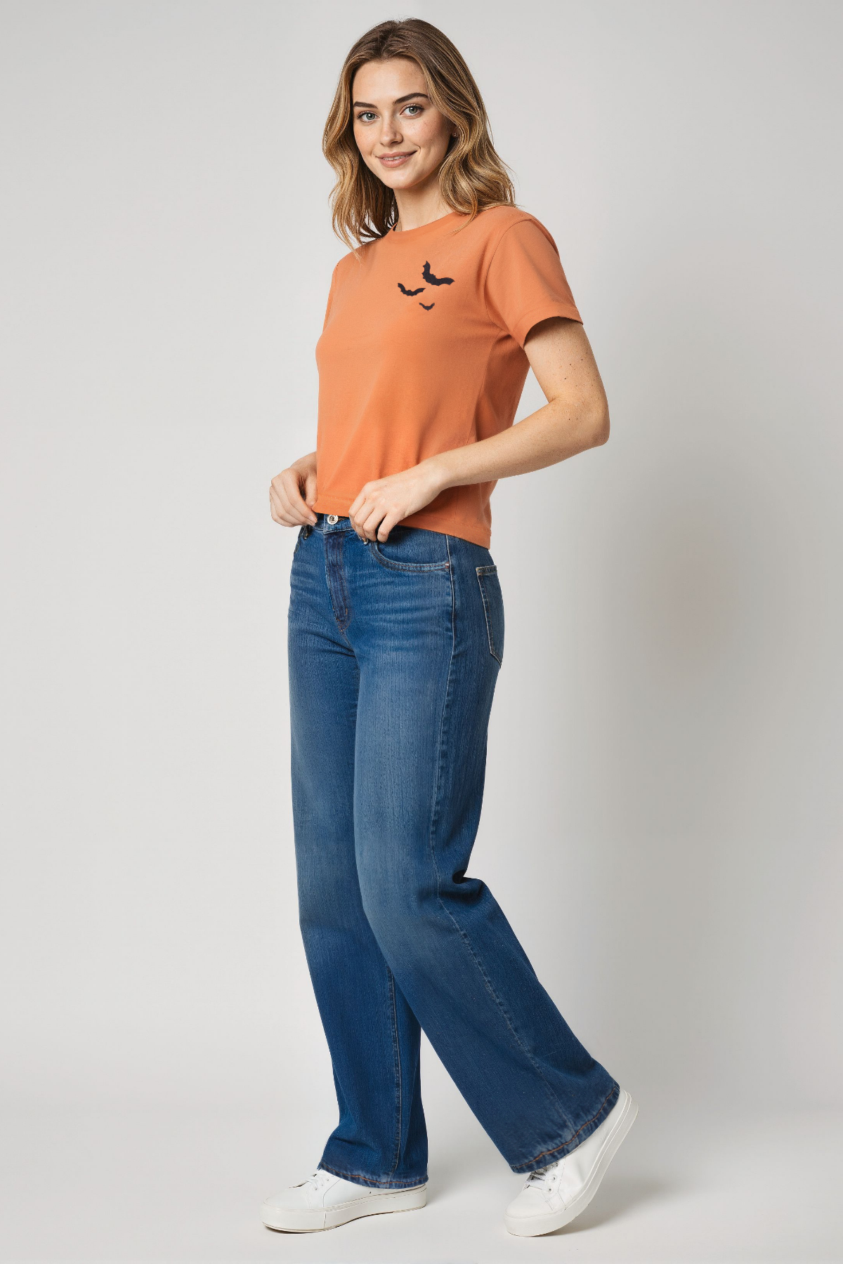 Woman wearing an orange t-shirt a 3 black bats design and blue jeans on a plain background