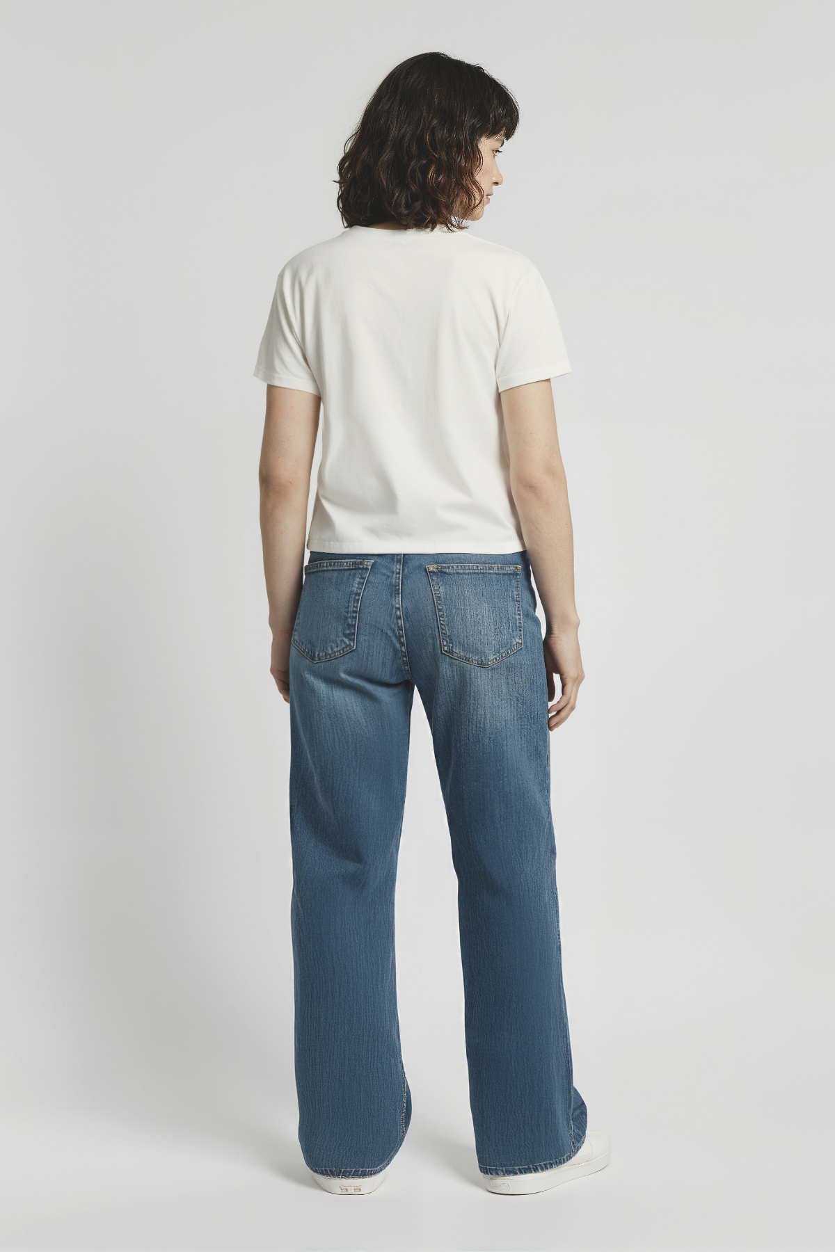 Back of woman wearing a white t-shirt and blue jeans on a plain background