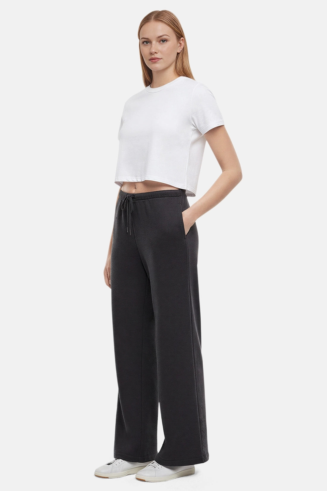 Side of woman wearing a white cropped t-shirt and heather black wide-leg drawstring sweatpants with hand in pocket on a white background