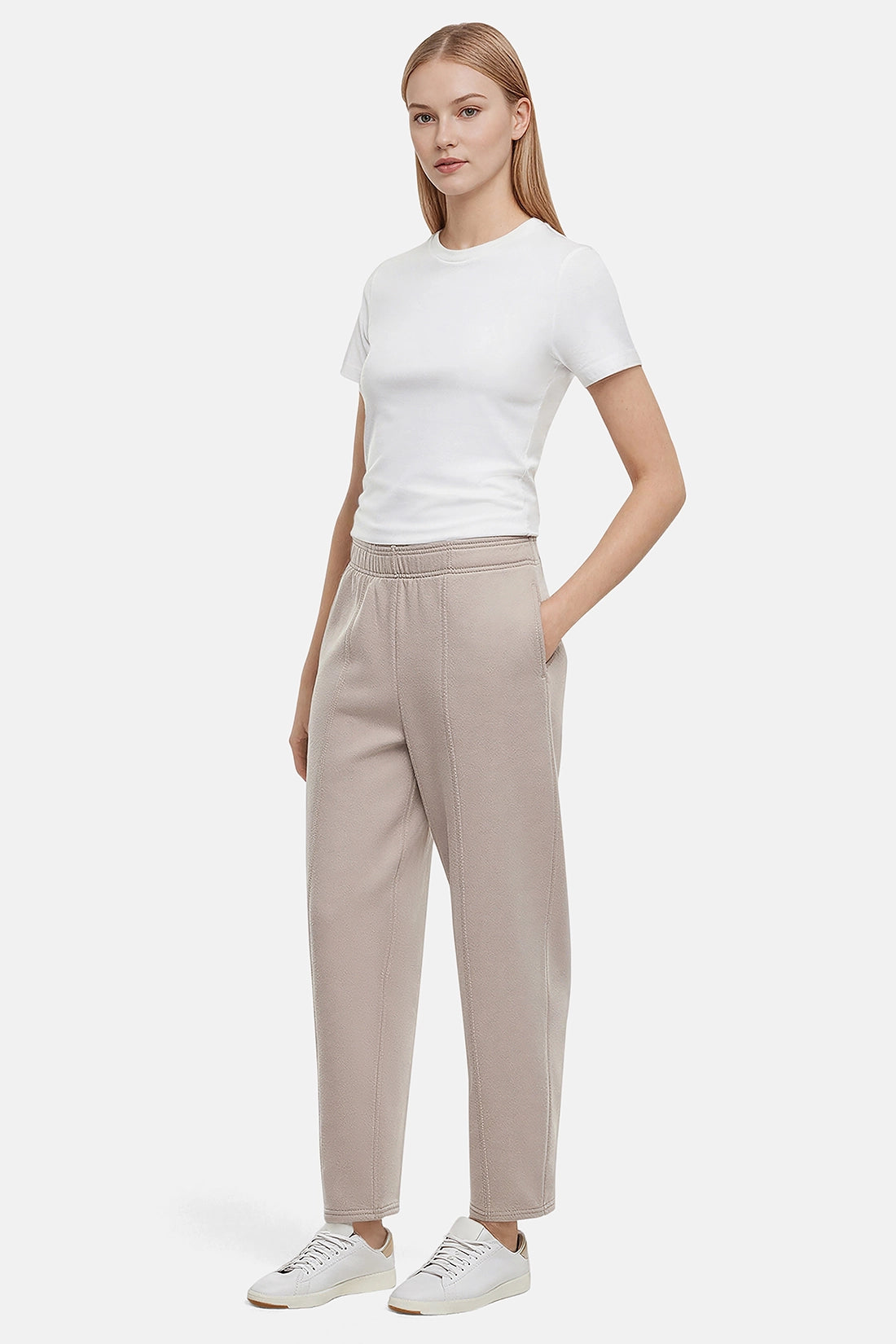 Woman wearing a white t-shirt and beige tapered leg pants with hand in pocket on a white background