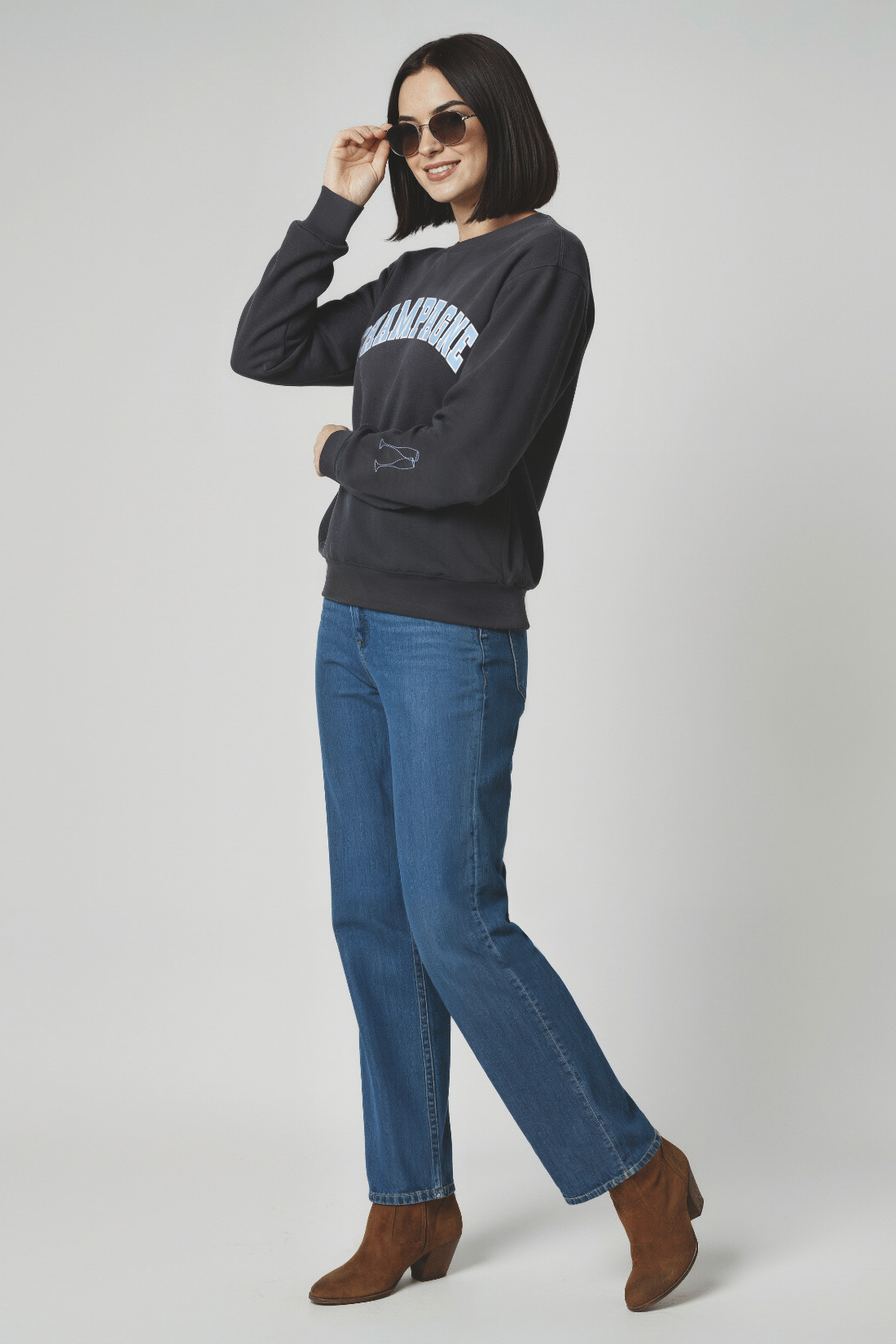 Woman wearing a dark gray sweatshirt with Champagne text, blue jeans, and brown boots on a white background