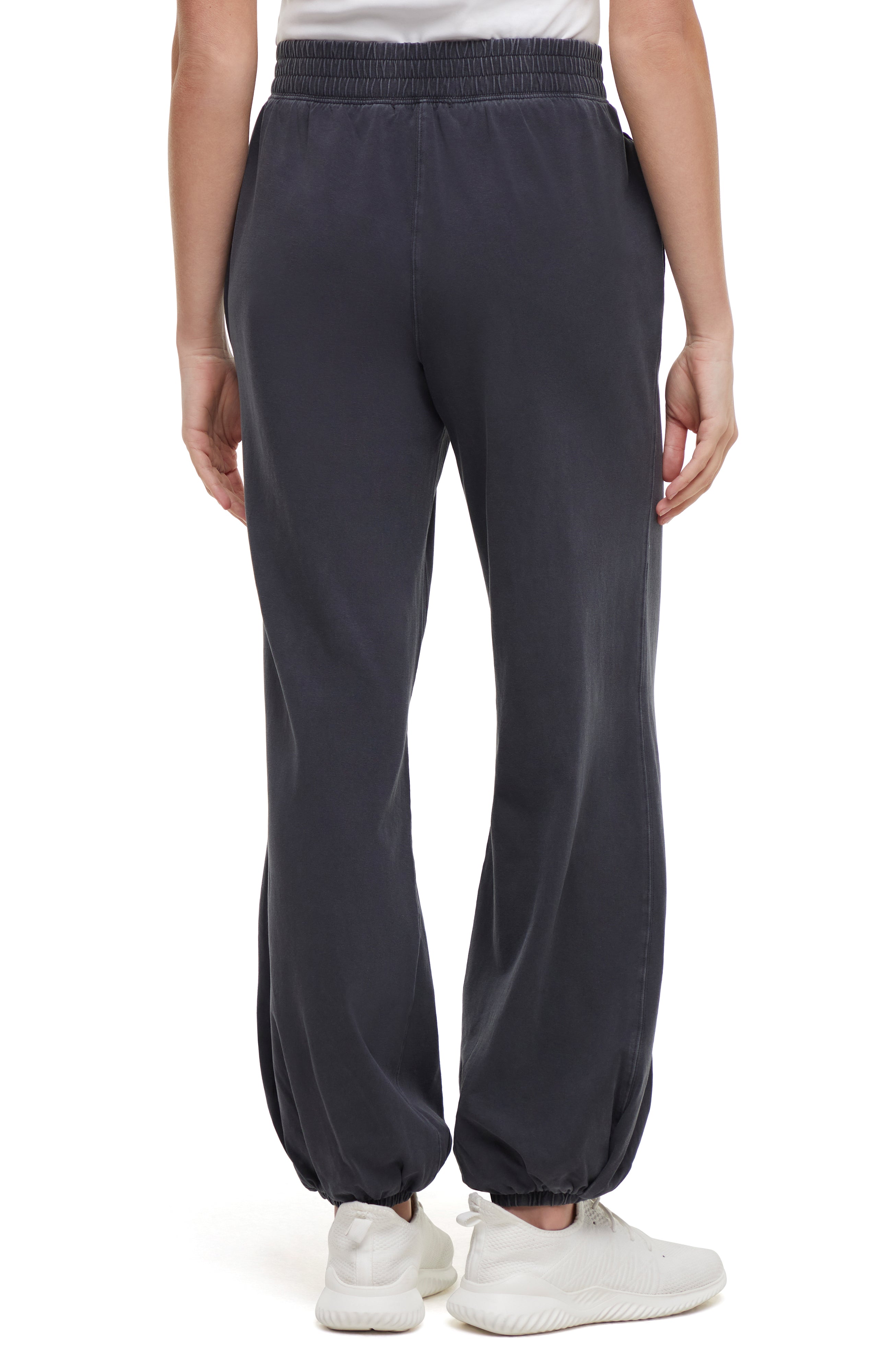 Back view of woman wearing dark gray pants and white sneakers on a white background.