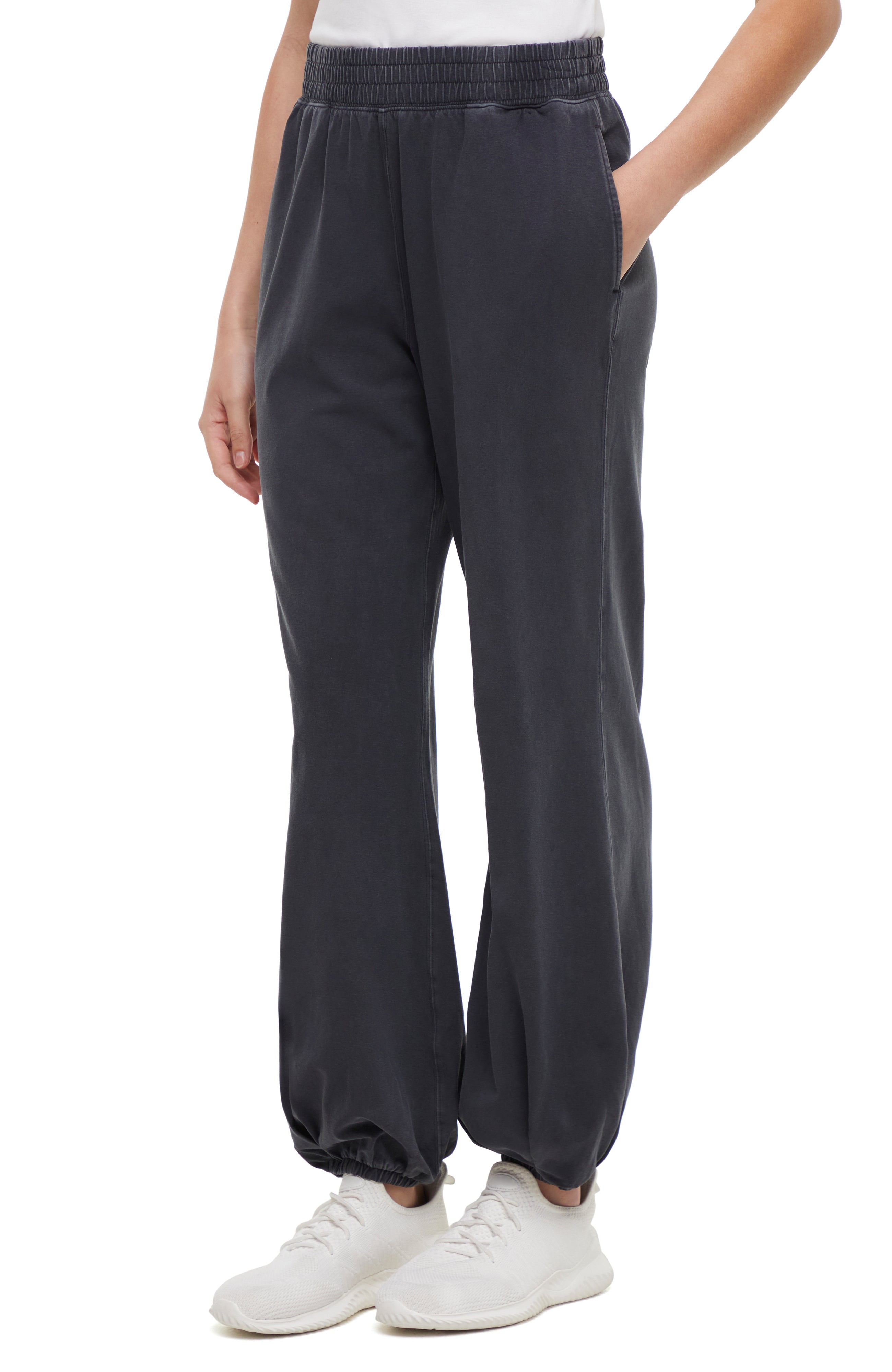 Front view of woman wearing dark gray pants and white sneakers on a white background.