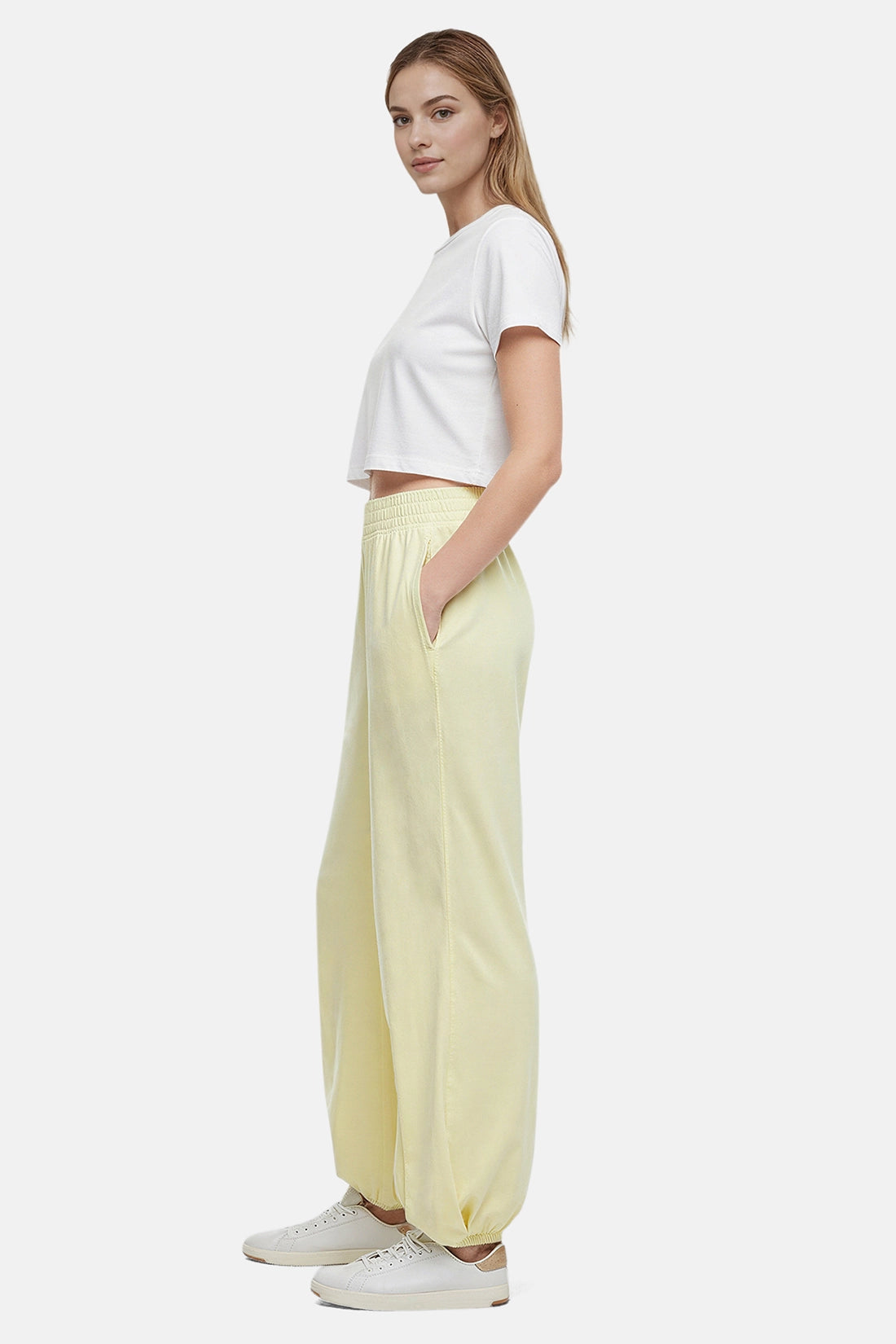 Side view of woman wearing yellow pants, white tee and white sneakers on a white background.