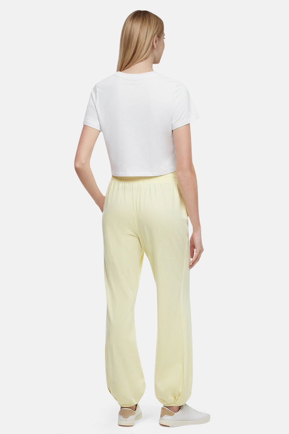 Back view of woman wearing yellow pants, white tee and white sneakers on a white background.
