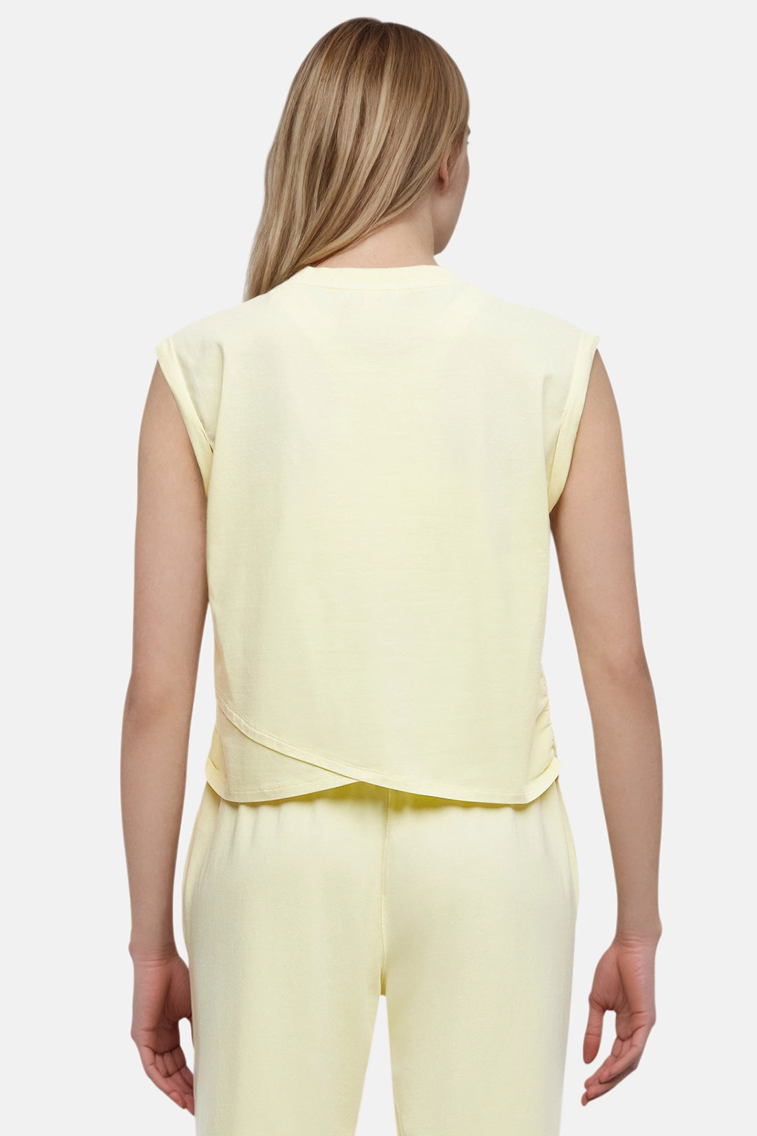 Back view of woman wearing a light-yellow sleeveless asymmetrical top and pants on a white background.