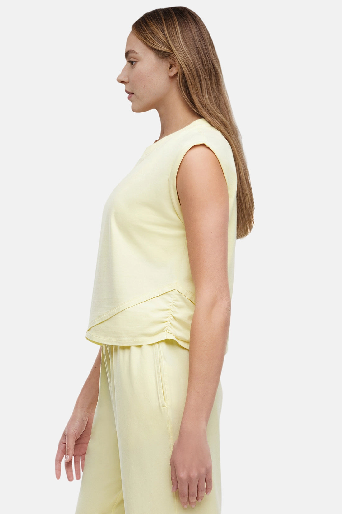Side view of woman wearing a light-yellow sleeveless asymmetrical top and pants on a white background.