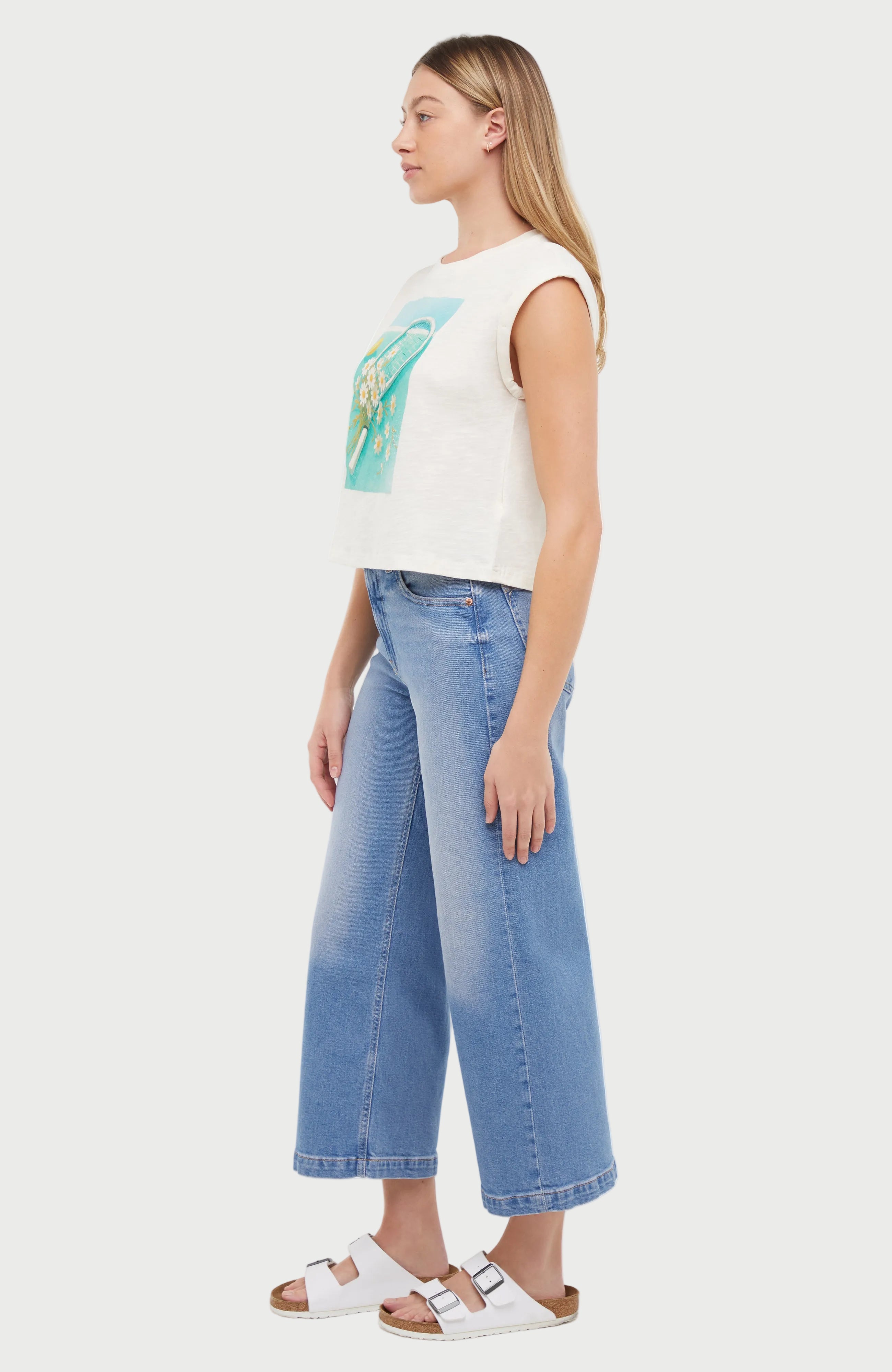 Side view of a woman wearing a white sleeveless shirt with a tennis racket and flowers design, standing against a white background.