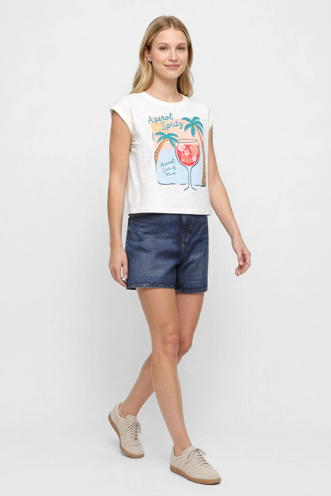 Side view of woman wearing white t-shirt with "Aperol Spritz; Aperol State of Mind" text and graphic design featuring a cocktail and palm trees and jean shorts on a white background.