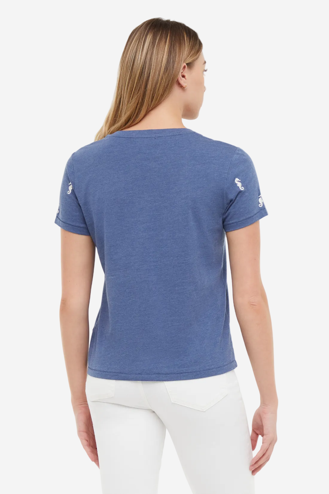 Back view of woman wearing a blue t-shirt with white embroidered seahorse patterns on sleeves and white jeans on a white background