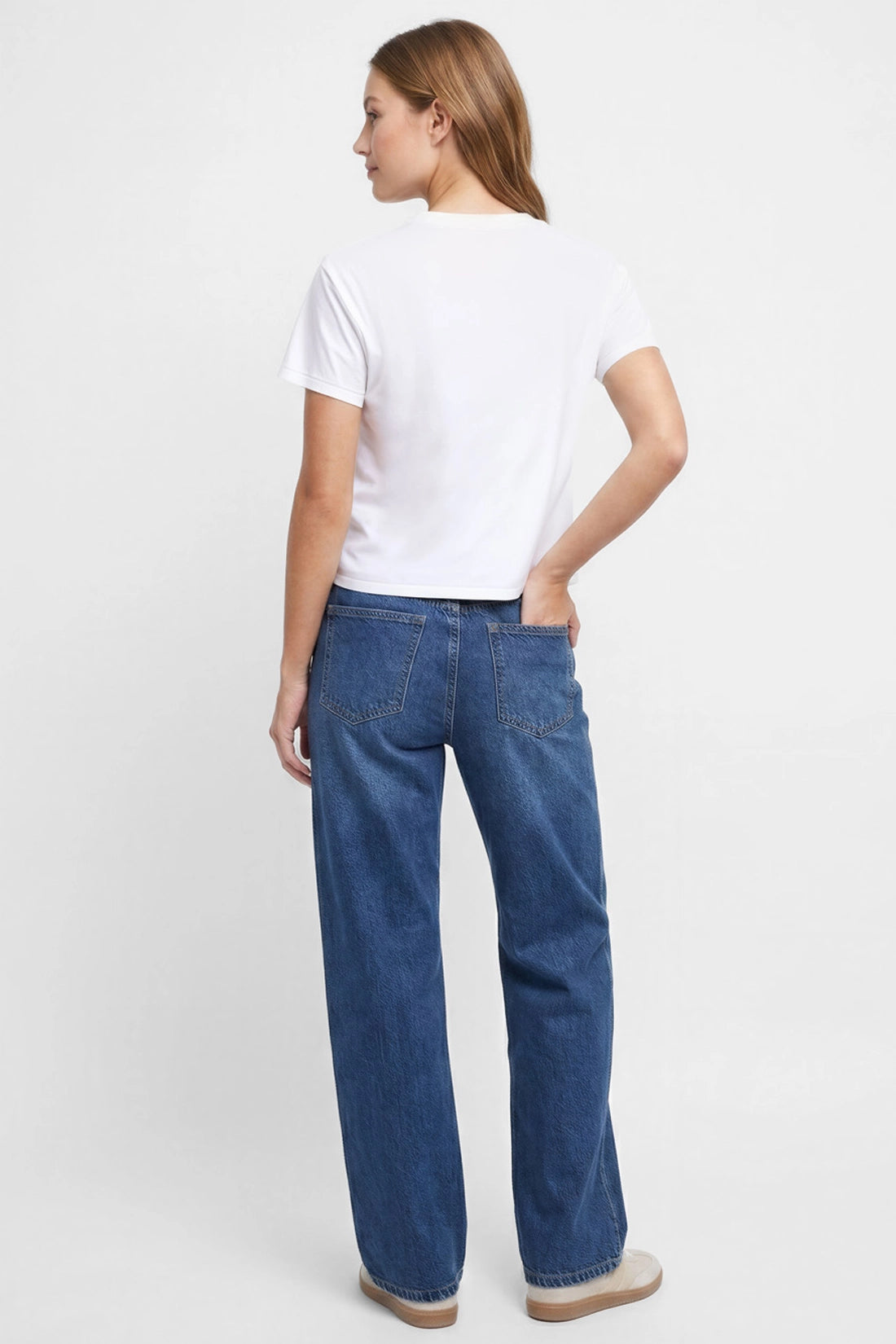 Back view of woman wearing a white t-shirt and blue jeans on a plain background.