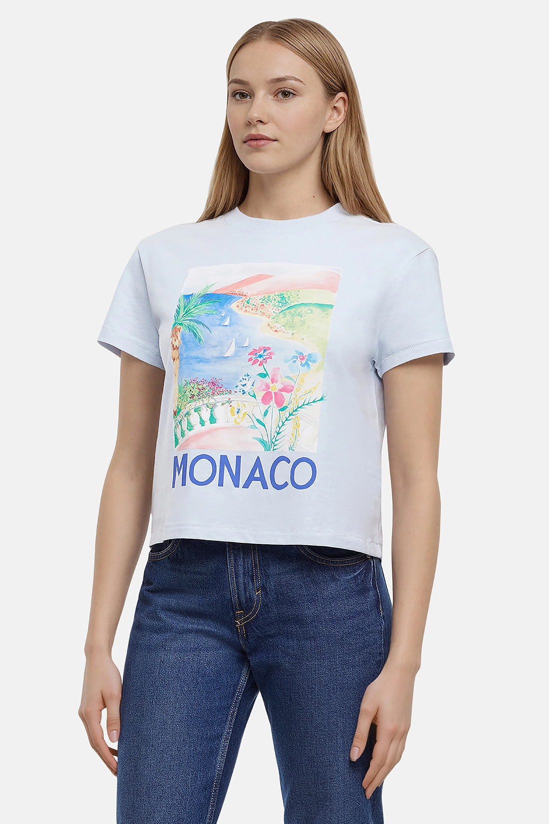 Woman wearing a light blue t-shirt with colorful beach, flowers, & sailboats graphic of Monaco and the &