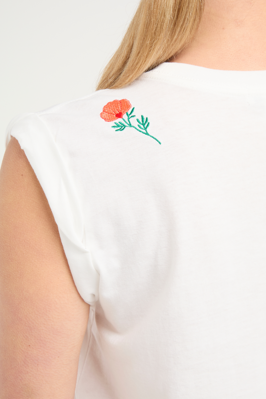 Close up back view of woman wearing white t-shirt with floral embroidery graphic design on top left shoulder and blue jeans on a white background.