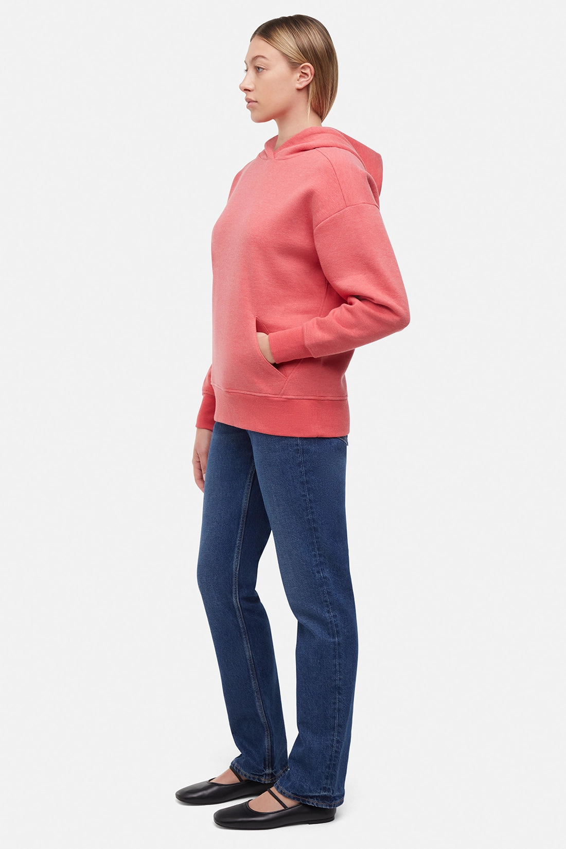 Side view of woman wearing a pink/red hoodie and blue jeans on a white background with hand in front kangaroo pocket.