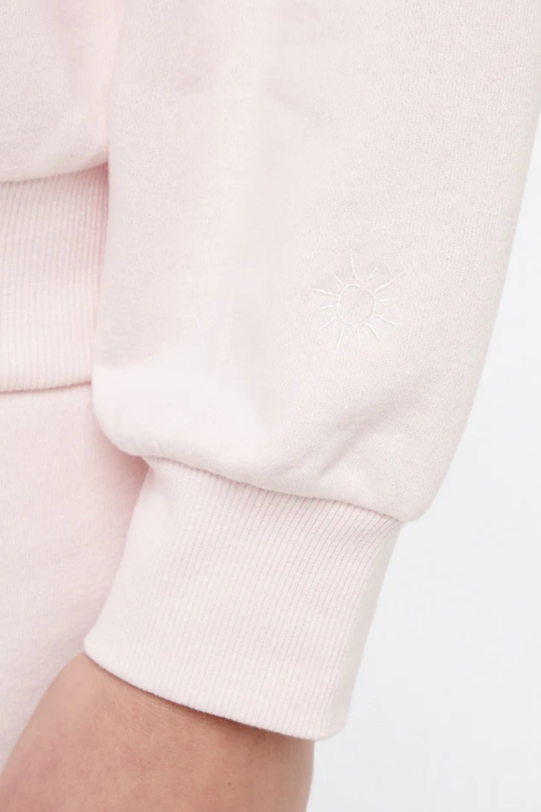 Close up front view of woman wearing a light pink sweatshirt with white sun on left sleeve hem on a white background.