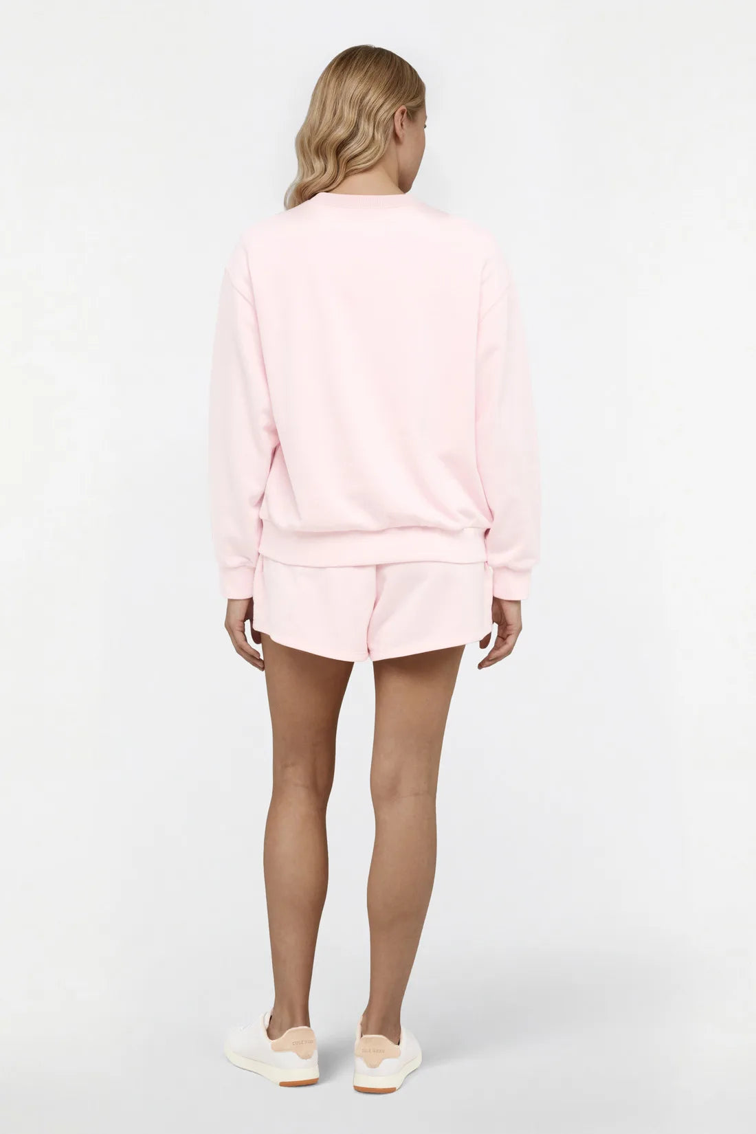 Back view of woman wearing a light pink sweatshirt and shorts set on a white background.