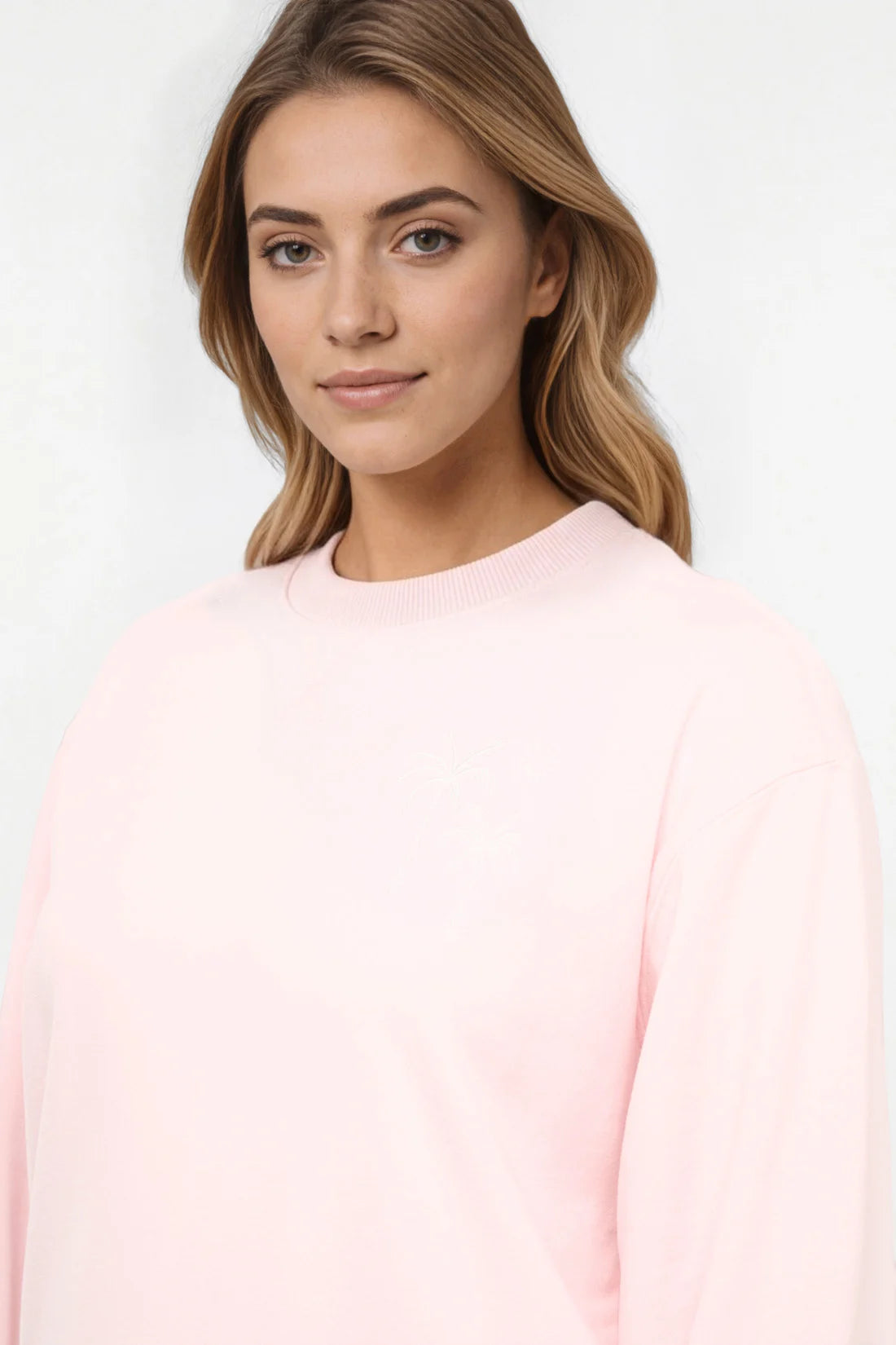 Close up front view of woman wearing a light pink sweatshirt with white palm tree on left chest on a white background.