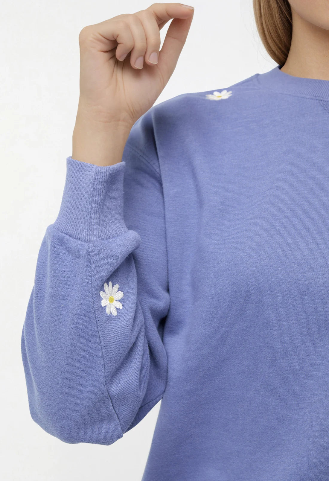 Close-up of a woman wearing a blue sweatshirt with daisy flower designs on shoulder and sleeve on a white background.
