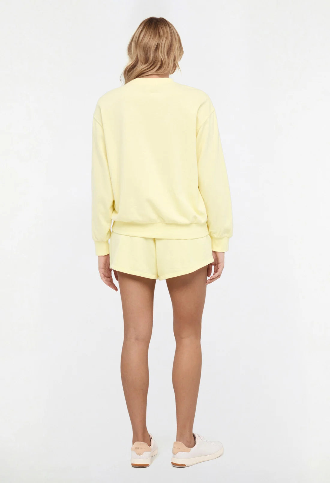 Back view of woman wearing a yellow sweatshirt and shorts on a light gray background.