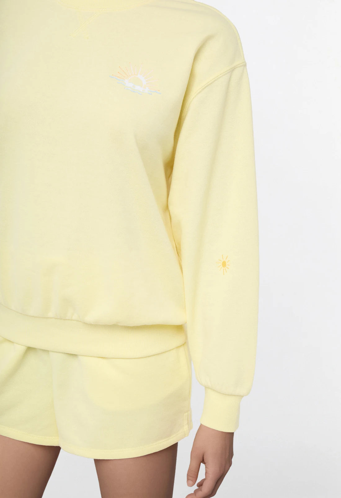 Close up sleeve view of woman wearing a yellow sweatshirt and shorts on a light gray background.
