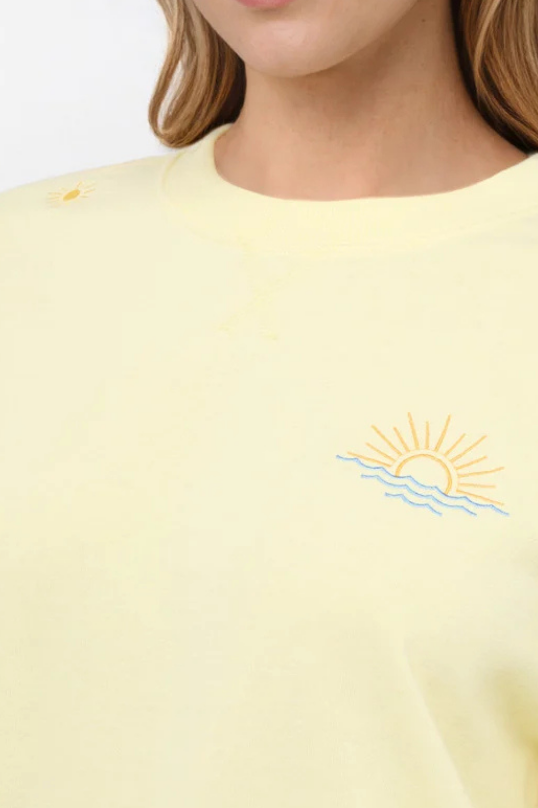 Close up view of woman wearing a yellow sweatshirt with sunset beach graphic on a light gray background.