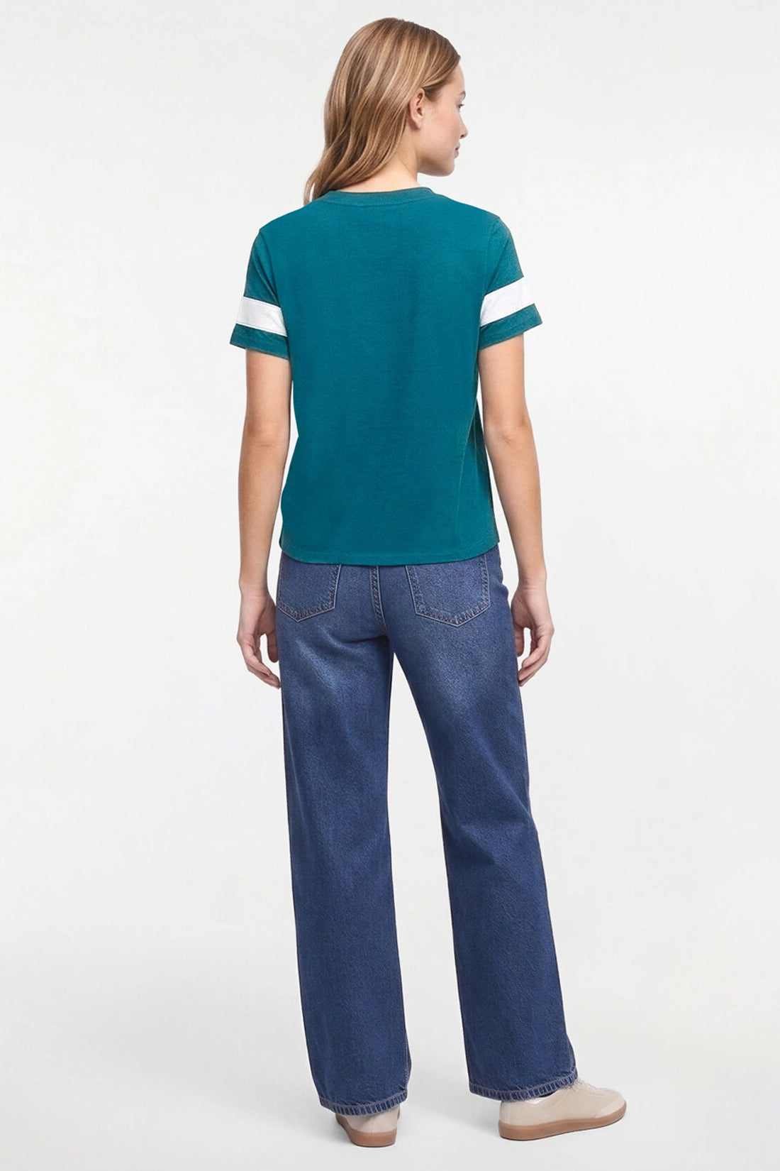 A woman viewed from the back wearing a teal short-sleeve t-shirt with white stripes on the sleeves and blue jeans, against a plain white studio background.