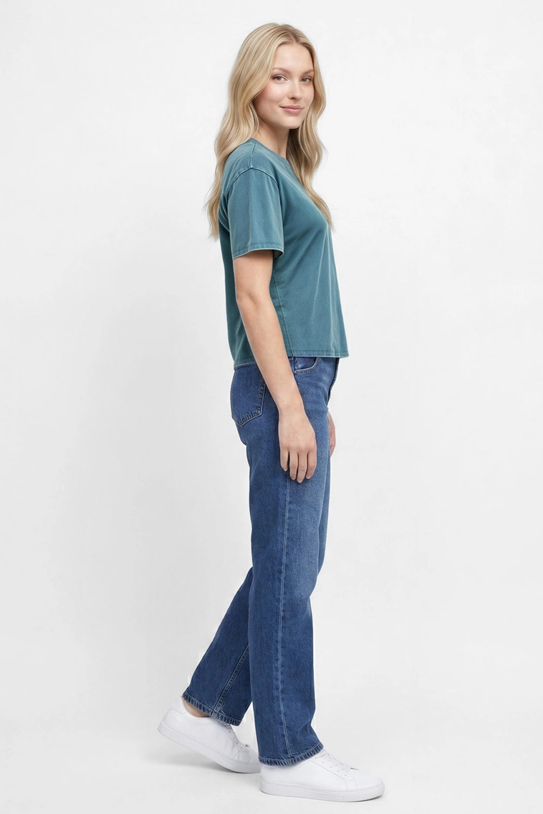Side view of woman wearing a teal green t-shirt and blue jeans on a white background.