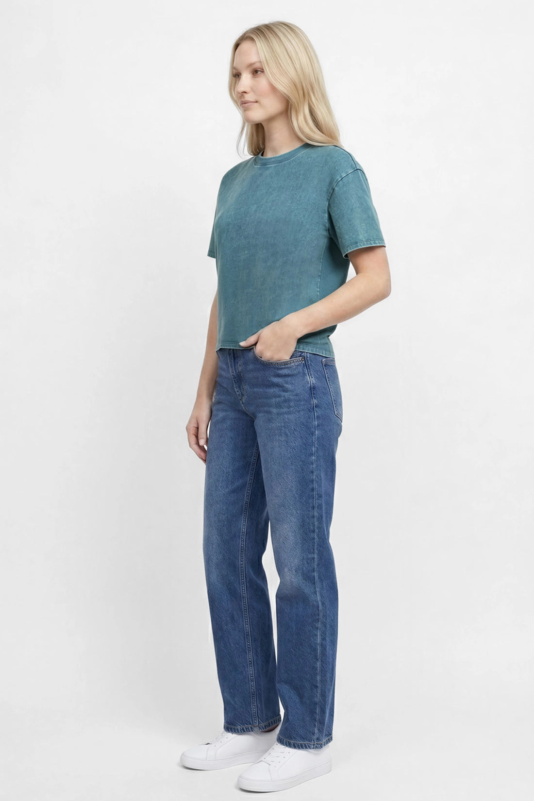 Side view of woman wearing a teal green t-shirt and blue jeans on a white background.