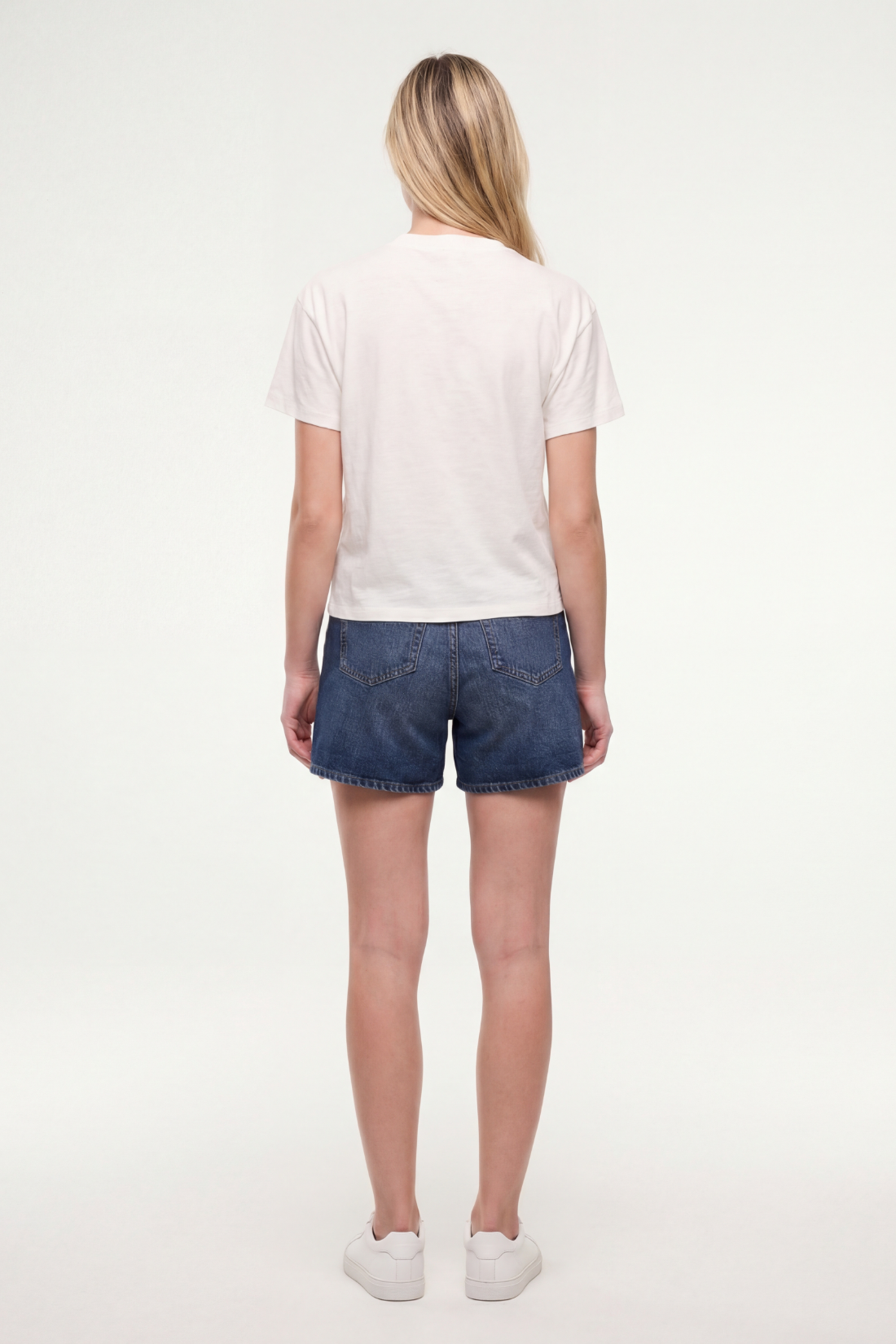 Back view of woman wearing a white t-shirt and jean shorts on a white background.