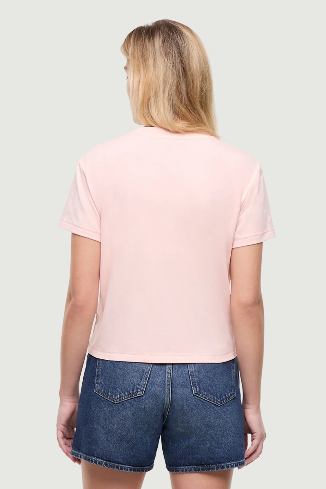 Back view of woman wearing a light pink t-shirt and denim shorts on a white background.
