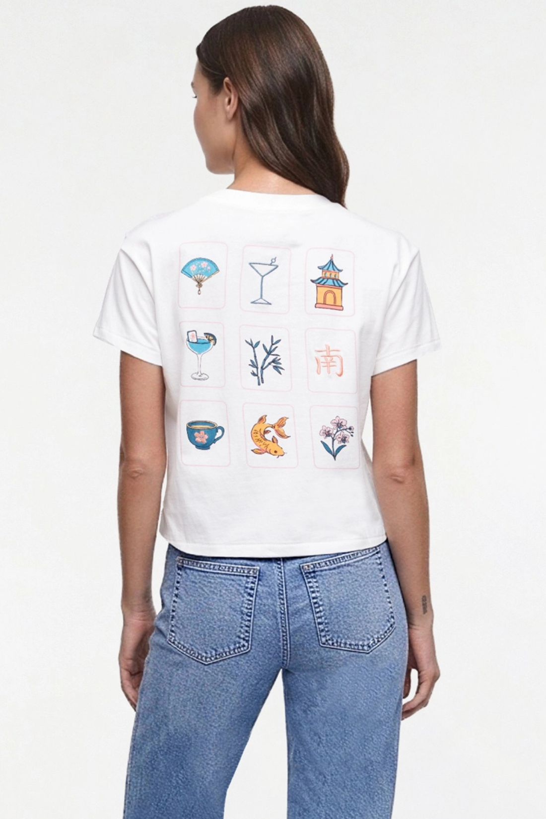 A woman viewed from the back wearing a white short-sleeve crop top T-shirt with a Mahjong grid of colorful cocktail-themed illustrations on the back, paired with blue jeans, against a plain white studio background.