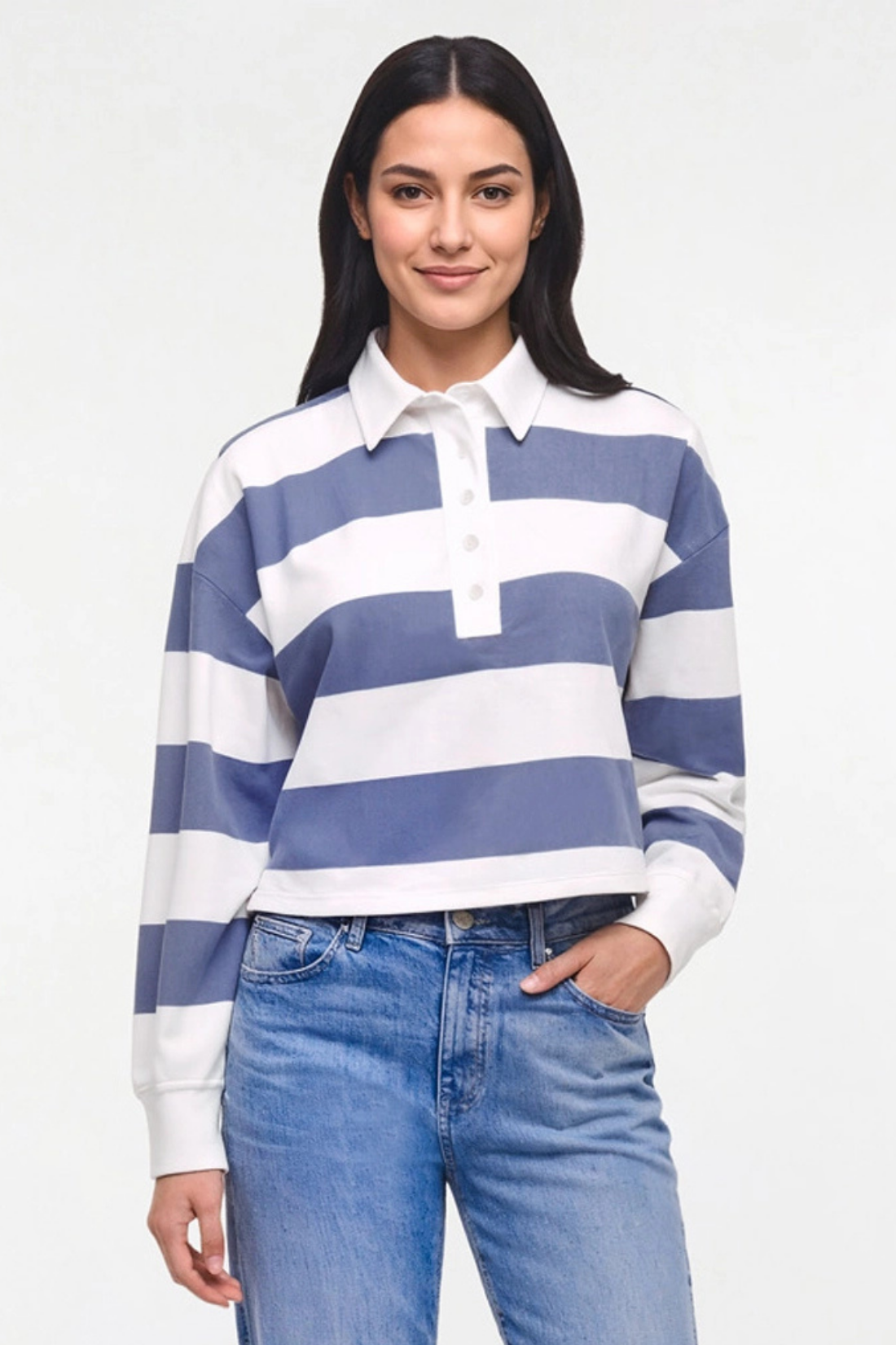 A woman modeling a long-sleeve rugby-style polo shirt with horizontal blue and white stripes, paired with light-wash blue jeans, against a plain white studio background.