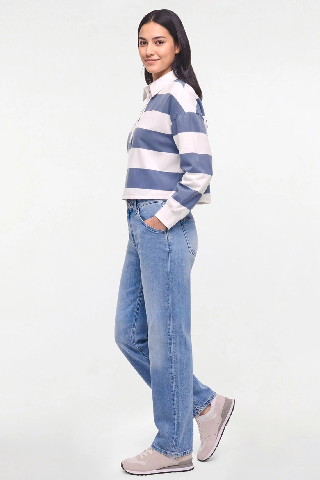 A woman modeling a blue and white striped long-sleeve rugby shirt with light-wash blue jeans and light gray sneakers, standing against a plain white studio background.