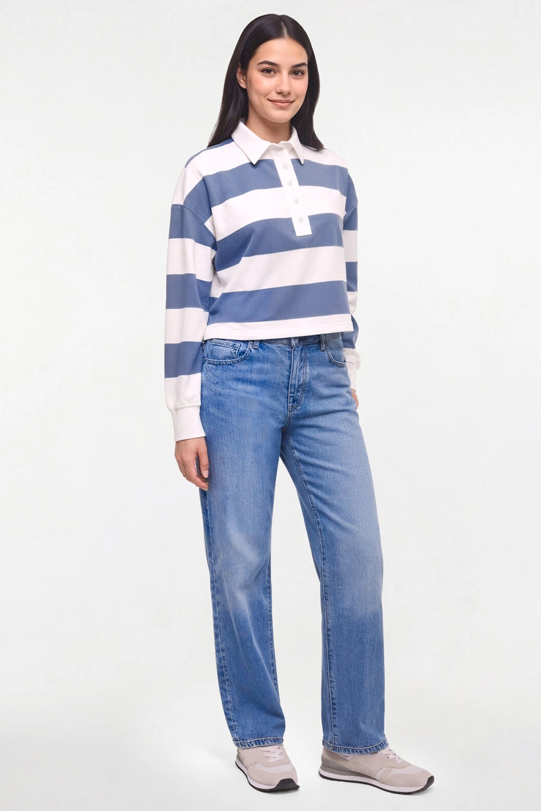 A woman modeling a blue and white striped long-sleeve rugby shirt with light-wash blue jeans and gray sneakers, standing against a plain white studio background.