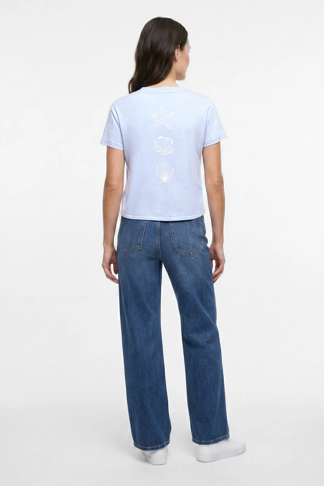 Back view of woman wearing light blue t-shirt with white starfish, shell, and pearl designs and blue jeans on a plain background.