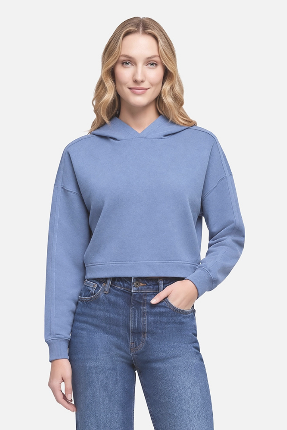A woman wearing a cropped, long-sleeve blue hoodie with a relaxed fit and drawstring-free hood, paired with blue jeans, against a light gray studio background.