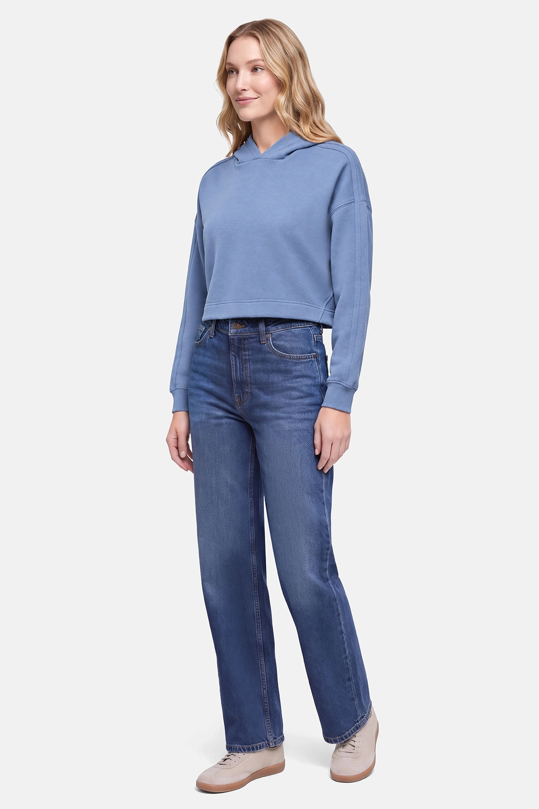 A woman modeling a light blue cropped hoodie and dark blue straight-leg jeans with beige sneakers, standing against a light gray studio background.
