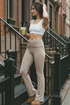 Woman in athletic wear white sports bra and tan yoga pants standing on a city street, holding a green drink.