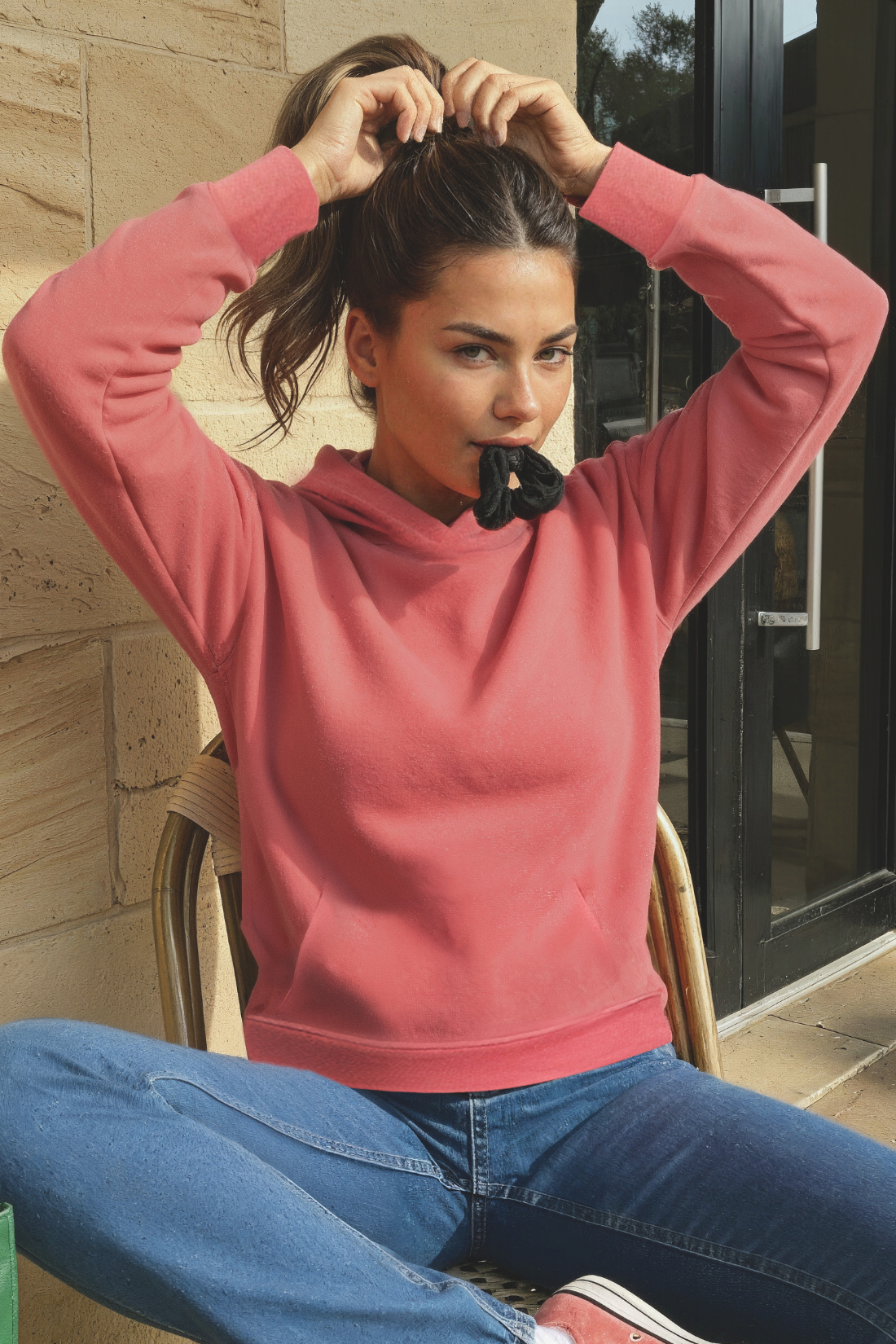 Woman in a pink hoodie and blue jeans sitting on a chair outdoors.