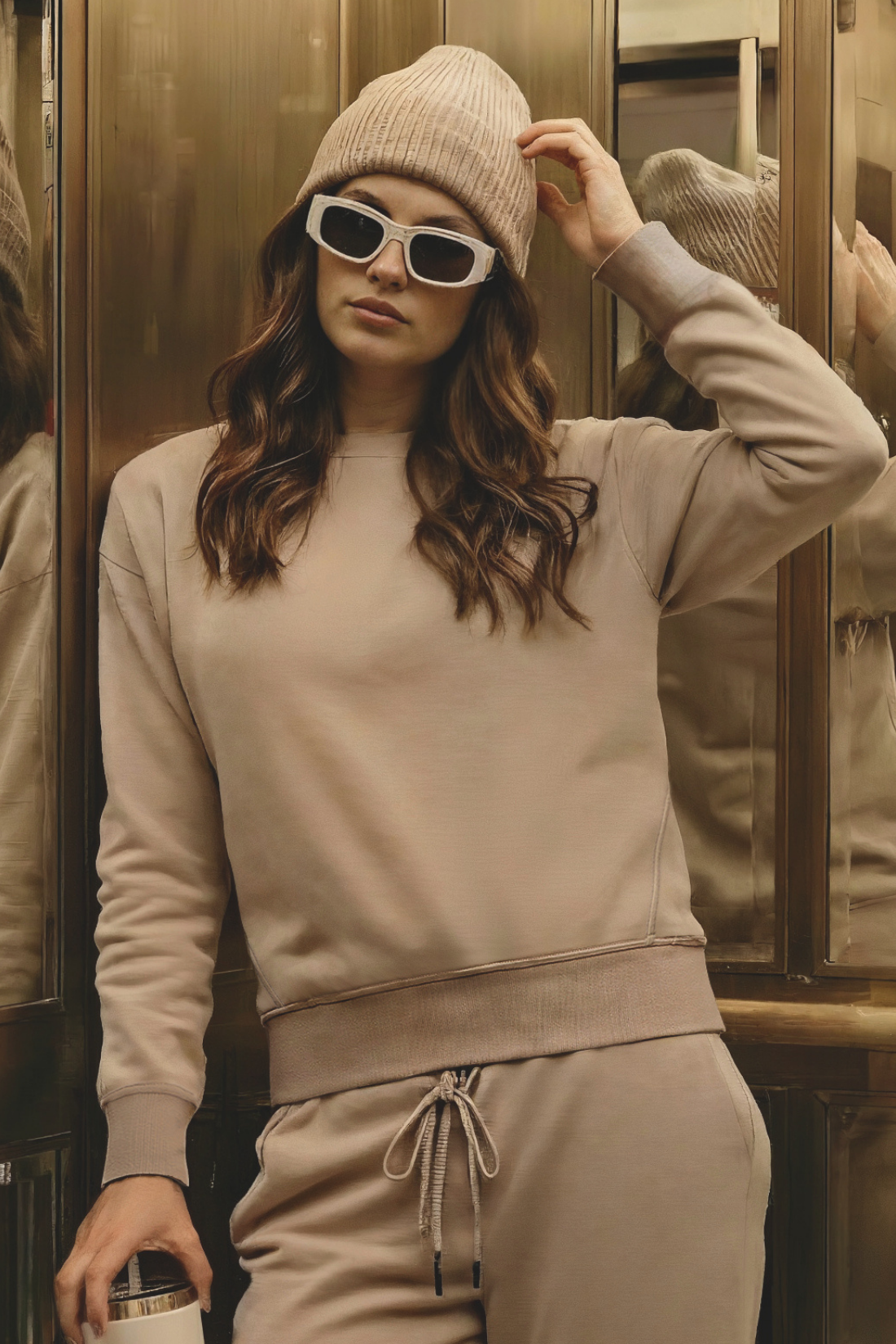 Woman in beige sweatshirt and sweatpants outfit with sunglasses standing in a reflective setting