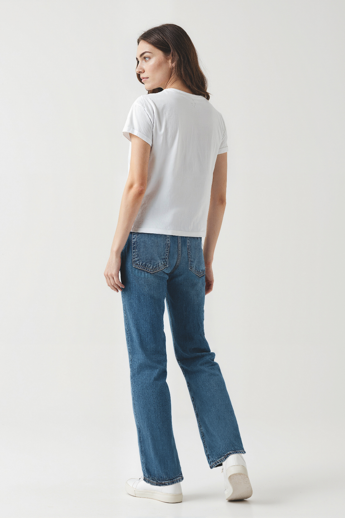Back of woman wearing a white t-shirt and blue jeans on a plain background.