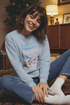 Woman wearing a light blue sweatshirt with a mountain graphic sitting on the floor in a cozy room.