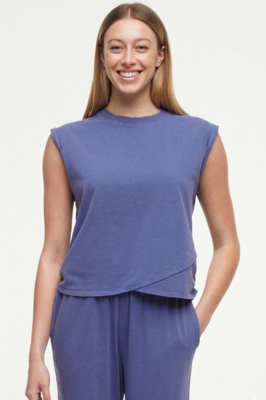 Front view of woman wearing a gray-blue sleeveless asymmetrical top and pants on a white background. 